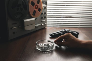 Özel ajan bobin kayıt cihazını dinliyor. Memur sigara içiyor. KGB konuşmaları gözetliyor. Kül tablasının yanında sigaralı bir el. Masada silah var..