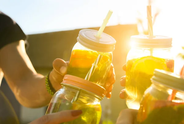 Friends holding fresh lemonade in jar with straw. Hipster summer party with drinks. Healthy vegan lifestyle. Eco-friendly in the nature. Lemons, oranges and berries with mint in the glass.