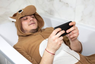 Kostümlü inek kostümü giymiş bir adam. Komik hayvan pijamalı adam banyoda uzanıyor ve cep telefonuyla oyun oynuyor..