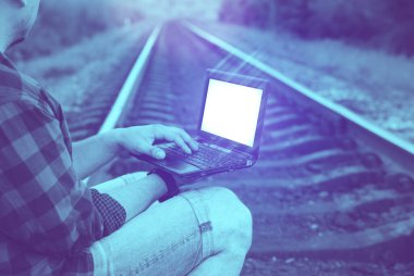 Dizüstü bilgisayarı olan adam rayların üzerinde oturuyor. Serbest hipster işçisi. İnsanoğlu seyahat ediyor..