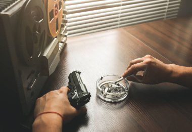Özel ajan bobin kayıt cihazını dinliyor. Memur sigara içiyor. KGB konuşmaları gözetliyor. Kül tablasının yanında sigaralı bir el. Masada silah var..