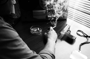 Özel ajan bobin kayıt cihazını dinliyor. Memur sigara içiyor. KGB konuşmaları gözetliyor. Kül tablasının yanında sigaralı bir el. Silahı masaya koy. Siyah beyaz fotoğraf.