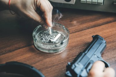 Özel ajan bobin kayıt cihazını dinliyor. Memur sigara içiyor. KGB konuşmaları gözetliyor. Kül tablasının yanında sigaralı bir el. Masada silah var..