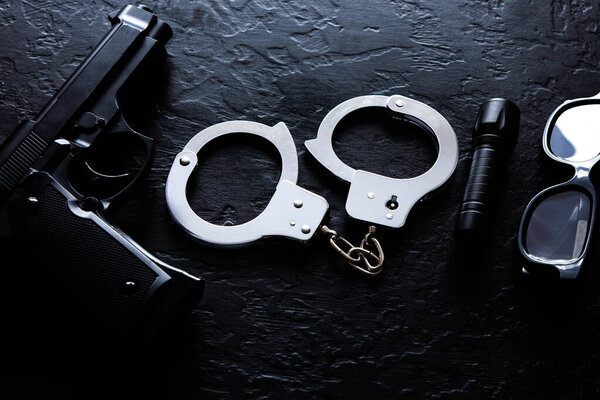 Black gun, flashlight, glasses and Police metal real handcuffs lie on the black background. Private detective work. Searching information. Crime and robbery, prison concept.