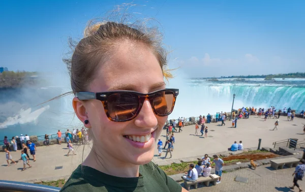 Radiant young girl happily smiling, wearing sunglasses, looking at camera, at Niagara Falls on sunny bright day. — Stock Photo, Image