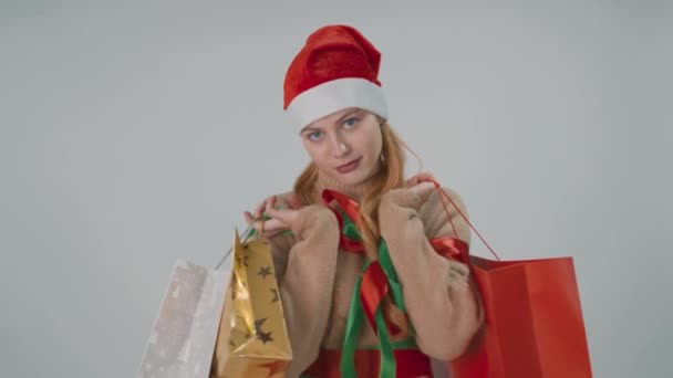 Szczęśliwa dziewczyna w kapeluszu Mikołaja, uśmiechnięty trzyma torby sprzedaży prezent izolowane na białym szarym. — Wideo stockowe