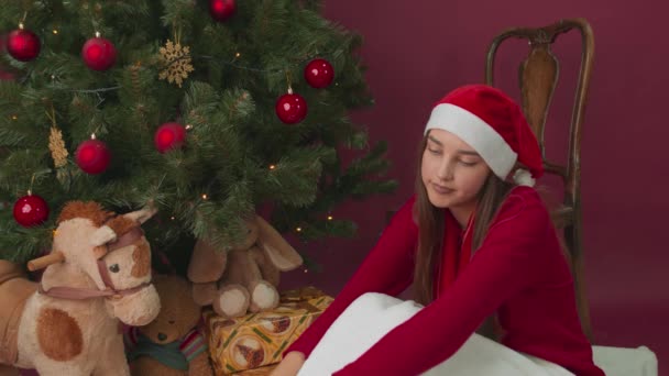 Uma menina triste em um chapéu de Papai Noel pela árvore sorriu depois de receber um presente. — Vídeo de Stock