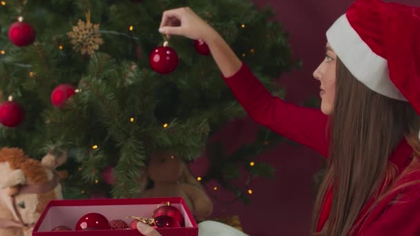 Dziewczyna ozdabia choinkę świątecznymi dekoracjami na czerwonym tle. — Wideo stockowe