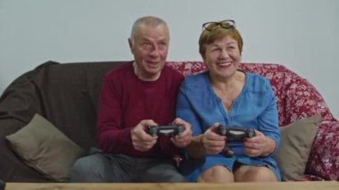 Yaşlı çift evde, kanepede joysticklerle oynuyor ve mutlu bir şekilde gülümsüyor..