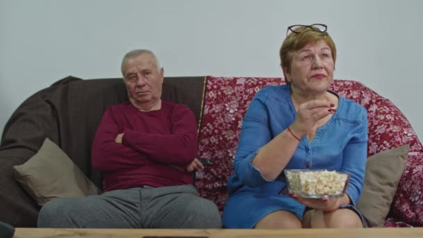 Starsze pary oglądają telewizję w domu na kanapie. Żona je popcorn.. — Wideo stockowe