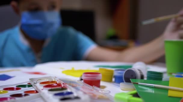 Kid w masce siedzi przy stole w pokoju i maluje obraz pędzlem z zieloną farbą — Wideo stockowe