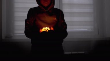 Hood 'lu Çocuğun Silueti. Karanlık bir odada yanan bir balkabağı mumunu tutan el.