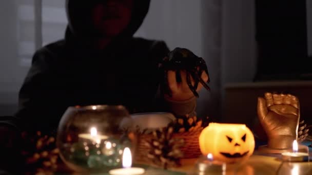 Mysteriöser Junge mit Kapuze spielt an Halloween mit Spinne bei Kerze im dunklen Raum — Stockvideo