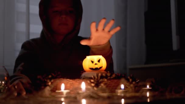Kluk v kápi vykouzlí dýňovou svíčku v tmavém halloweenském pokoji — Stock video