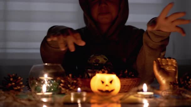 Schamanenjunge mit Kapuze führt Ritus bei Kerze im dunklen Raum an Halloween vor — Stockvideo