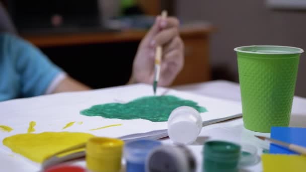 Мальчик в очках и голубой футболке рисует картину на бумаге с красками — стоковое видео