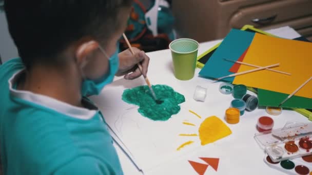 Парень в маске сидит за столом в комнате и рисует кистью с зеленой краской — стоковое видео