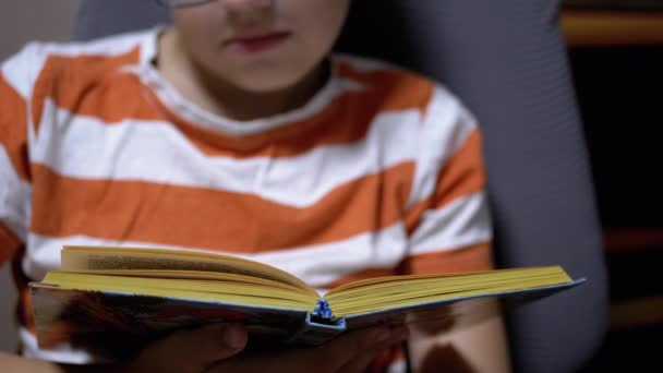 Серйозний хлопчик з окулярами читає цікаву книжку вдома — стокове відео