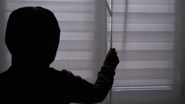 Silhouet van eenzame kind in kap zwaaiend met een gordijn, kijkend naar het raam. Donker — Stockvideo