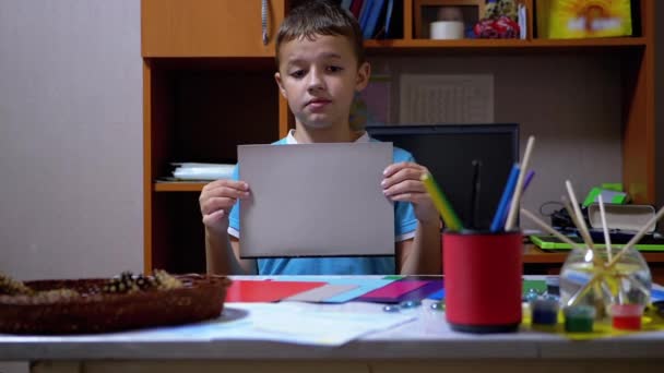 Серйозний хлопчик у блакитній футболці піднімає руки і показує сірий на кольоровому папері — стокове відео