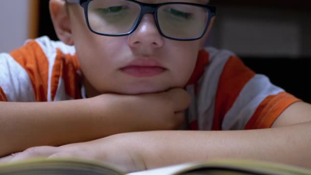 Inquisitivo, un chico serio con gafas está leyendo un libro interesante en casa — Vídeos de Stock