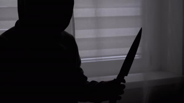 Silhouette di bambino solitario in cappuccio con coltello in mano vicino alla finestra. Adolescente assassino — Video Stock