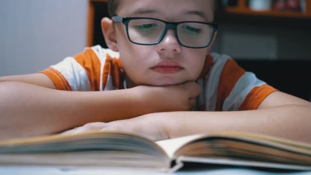 Inquisitivo, un chico serio con gafas está leyendo un libro interesante en casa — Vídeos de Stock