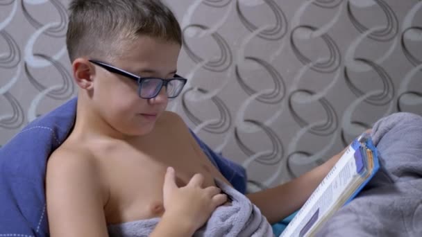 Ospalý chlapec s brýlemi čte zajímavou knihu, zívá na posteli v dece — Stock video