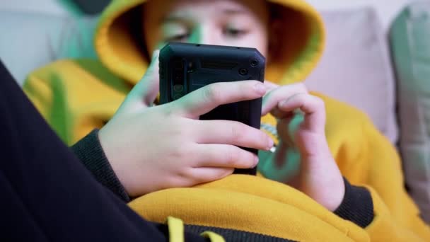 Pojke i gul Hoody sitter på soffan, ser meddelanden, spela på smartphone hemma — Stockvideo