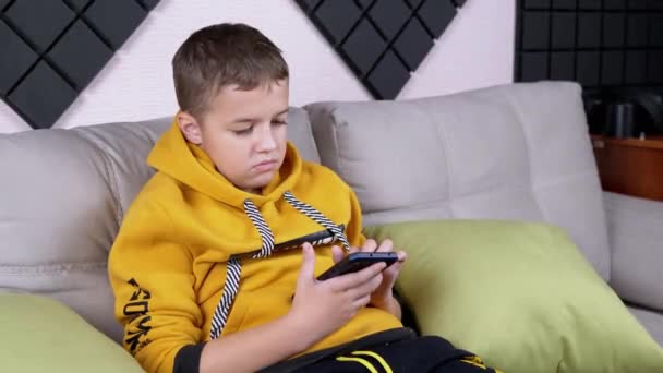 Nastolatek w żółtym kapturze siedzi na kanapie Gra mobilna na smartfonie w domu — Wideo stockowe