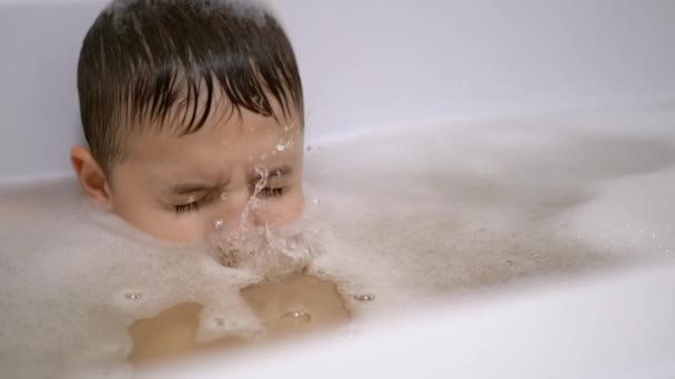 Junge badet in Schaumwasser in Badewanne, taucht unter Wasser, bläst Blasen aus nächster Nähe. — Stockvideo