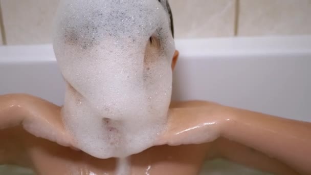 Мальчик купается в ванной, играет с пузырьками мыльной пены, делает смешные пенные маски — стоковое видео