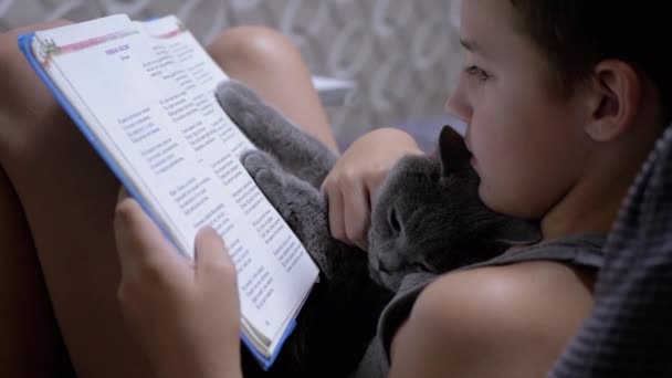 Sonríe chico con gafas leyendo un interesante libro con gato en las manos en casa — Vídeos de Stock