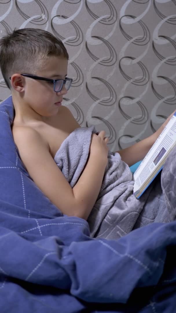 Seriöser Junge mit Brille liest ein interessantes Buch im Sitzen auf Bettdecke — Stockvideo
