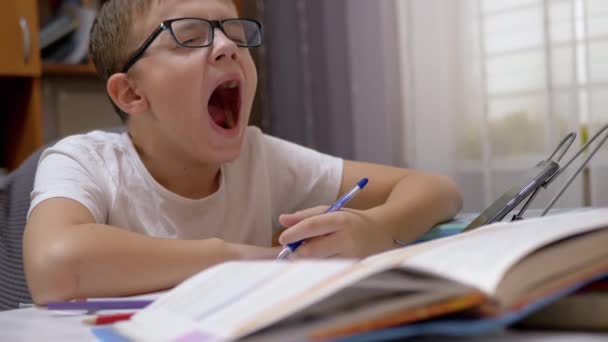 Уставший школьник в очках зевает, пишет ручкой в блокноте. 4K — стоковое видео