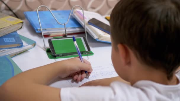 Школьник левша пишет с ручкой в ноутбуке делает домашнюю работу использовать смартфон — стоковое видео