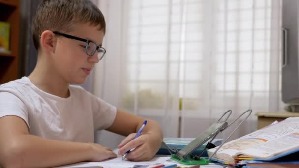 Estudante com óculos escreve com caneta no caderno, falando no Skype, acenando à mão — Vídeo de Stock