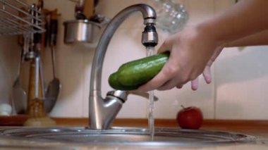 Modern bir mutfaktaki musluktan akan suyla yeşil salatalık yıkayan kadın.