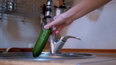 Modern bir mutfaktaki musluktan akan suyla yeşil salatalık yıkayan kadın.