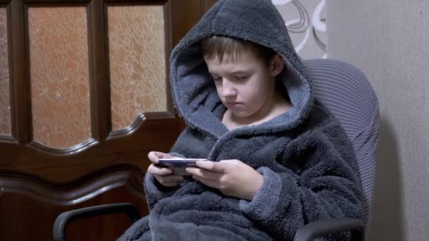 Chłopiec w szarym szlafroku siedzi na krześle Grając w grę mobilną na smartfonie w domu — Wideo stockowe