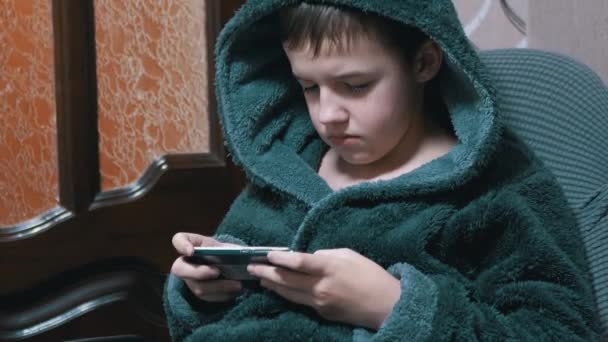 Jongen in een grijze badjas zit in stoel spelen Mobile Game op Smart Phone thuis — Stockvideo