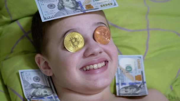 Sonriente adolescente miente, en una cama con dólares, Bitcoins de oro en los ojos. Humor. 4K — Vídeos de Stock