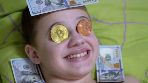 Улыбающийся мальчик лжет, на кровати с долларами, золотые биткоины на глазах. Юмор. Zoom — стоковое видео
