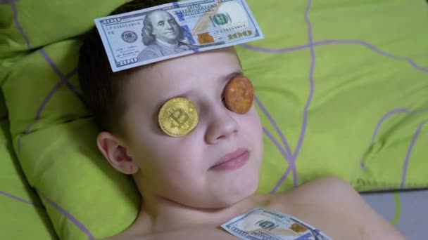 Uśmiechnięty nastolatek kłamie, na łóżku z dolarami, złote bitcoiny na oczach. Żarty. 4K — Wideo stockowe