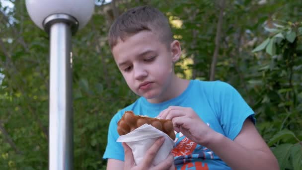 Criança senta-se no banco, come sorvete cremoso em uma taça de waffle, em um parque na rua — Vídeo de Stock