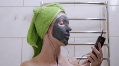 Yüzünde kil maskesi olan bir kız Smartphone 'da kulaklıkla müzik dinliyor.