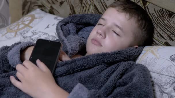 Зияющий ребенок в домашнем сером платье падает спать со смартфоном в руках — стоковое видео