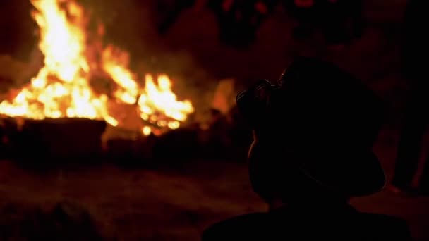 Maschio in un cappello si siede e guarda la fiamma luminosa di un falò notturno nella foresta — Video Stock