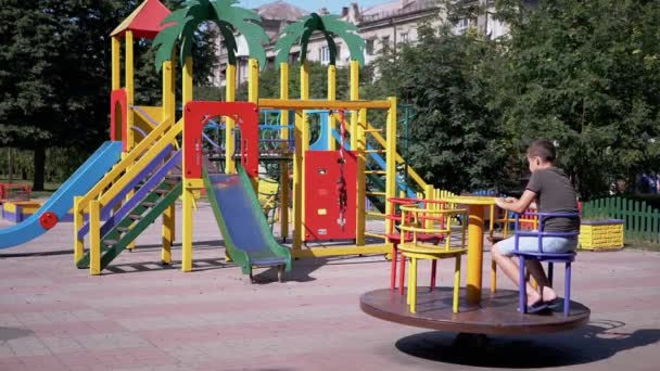 Одинокий грустный ребенок катается на пустой карусели во время карантина на детской площадке — стоковое видео
