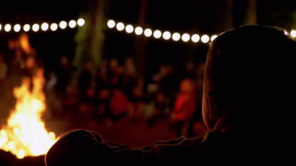 Ребенок сидит и смотрит на ночной костер в лесу с группой туристов. 4K — стоковое видео